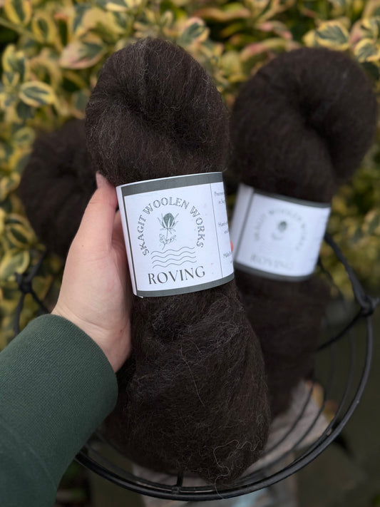Hand holding a hank of dark brown wool roving above a basket with more hanks, against a leafy background.