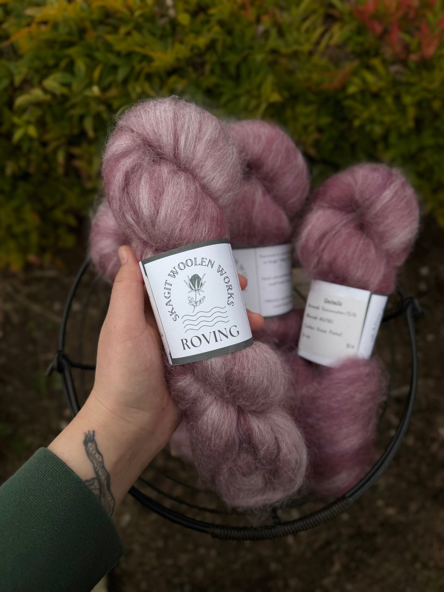 Hand holding a hank of dusty pink wool and silk roving with a label above a basket of roving and a blurred natural background.
