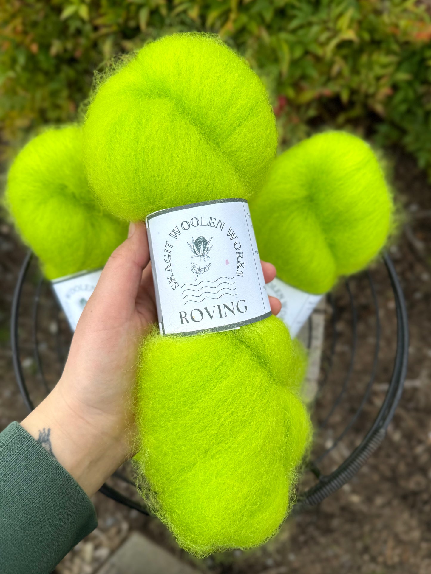 Hand holding a hank of neon green Coopworth wool roving with a label above a basket of roving and a blurred natural background.