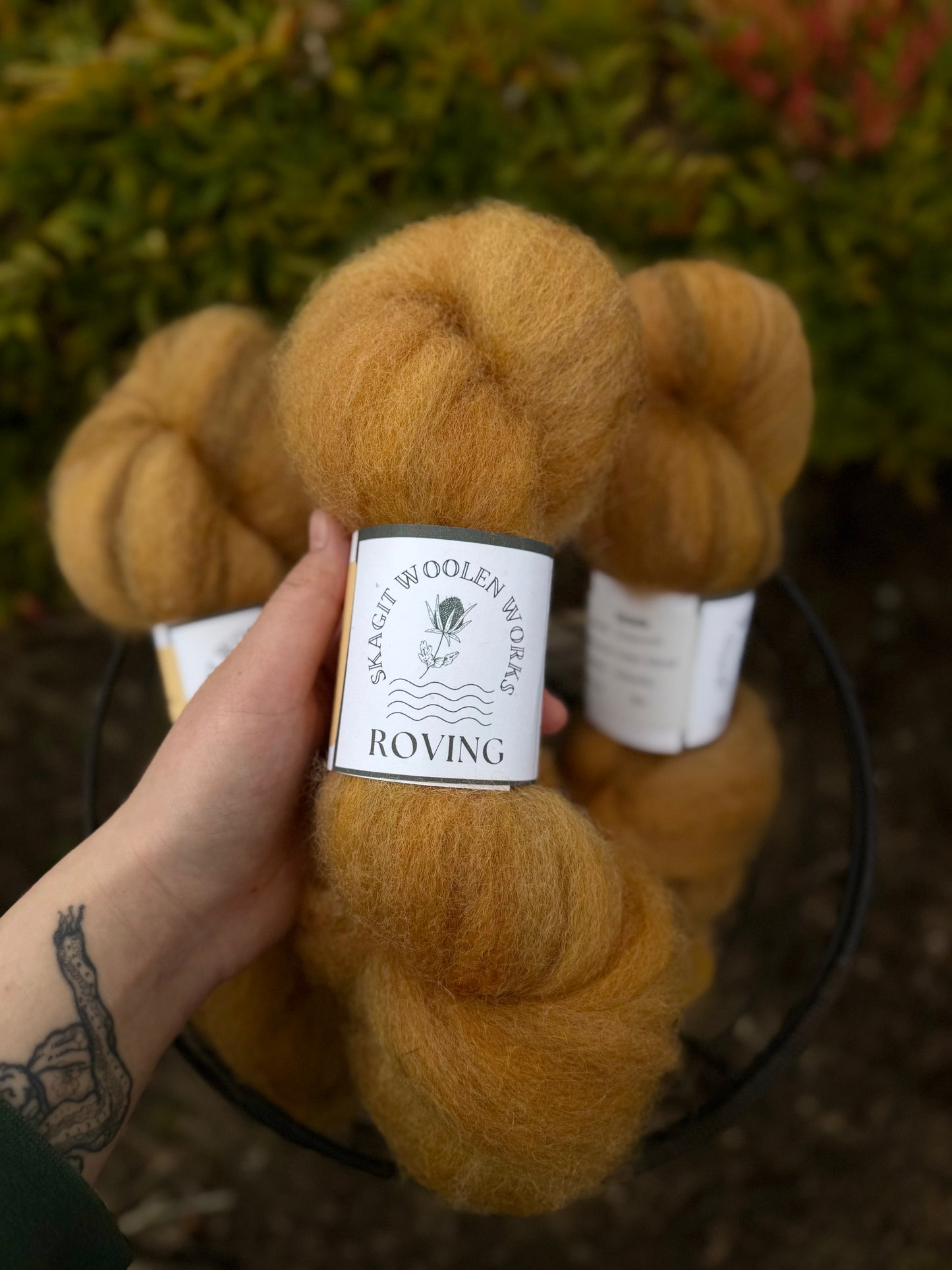 Hand holding a hank of orange Coopworth wool roving with a label above a basket of roving and a blurred natural background.
