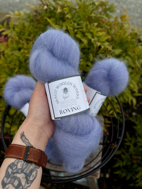 Hand holding a hank of vibrant periwinkle roving, above a basket of more hanks.