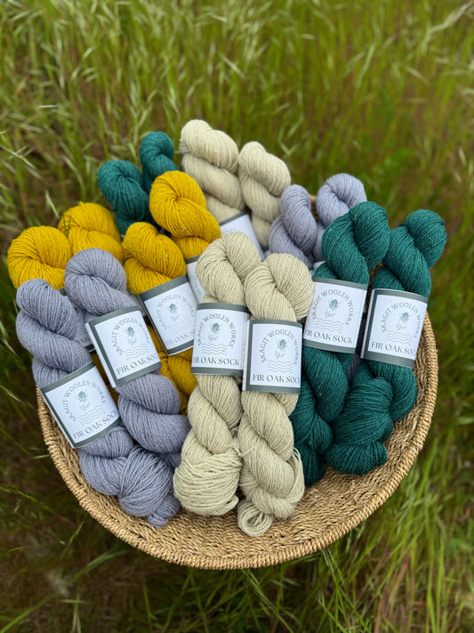Basket full of Romney Suffolk cross yarn, with 4 different colorways: Deep teal, muted periwinkle, a sunny yellow, and natural warm light grey.