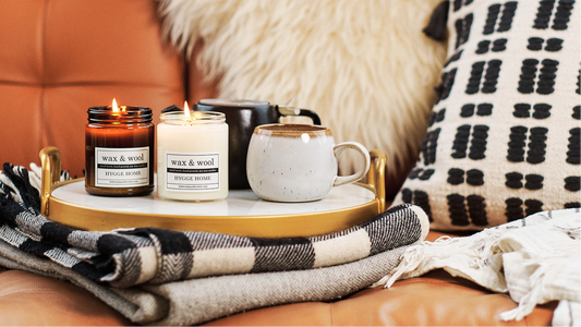 2 candles displayed on a tray with mugs, surrounded by blankets and pillows on a leather couch.